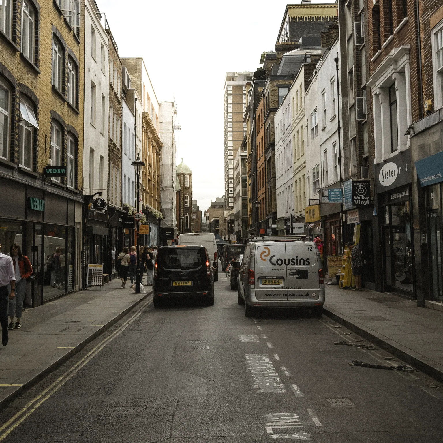 A view along Berwick Street with pedestrians, vans and shopfronts between rows of brick and white buildings.
