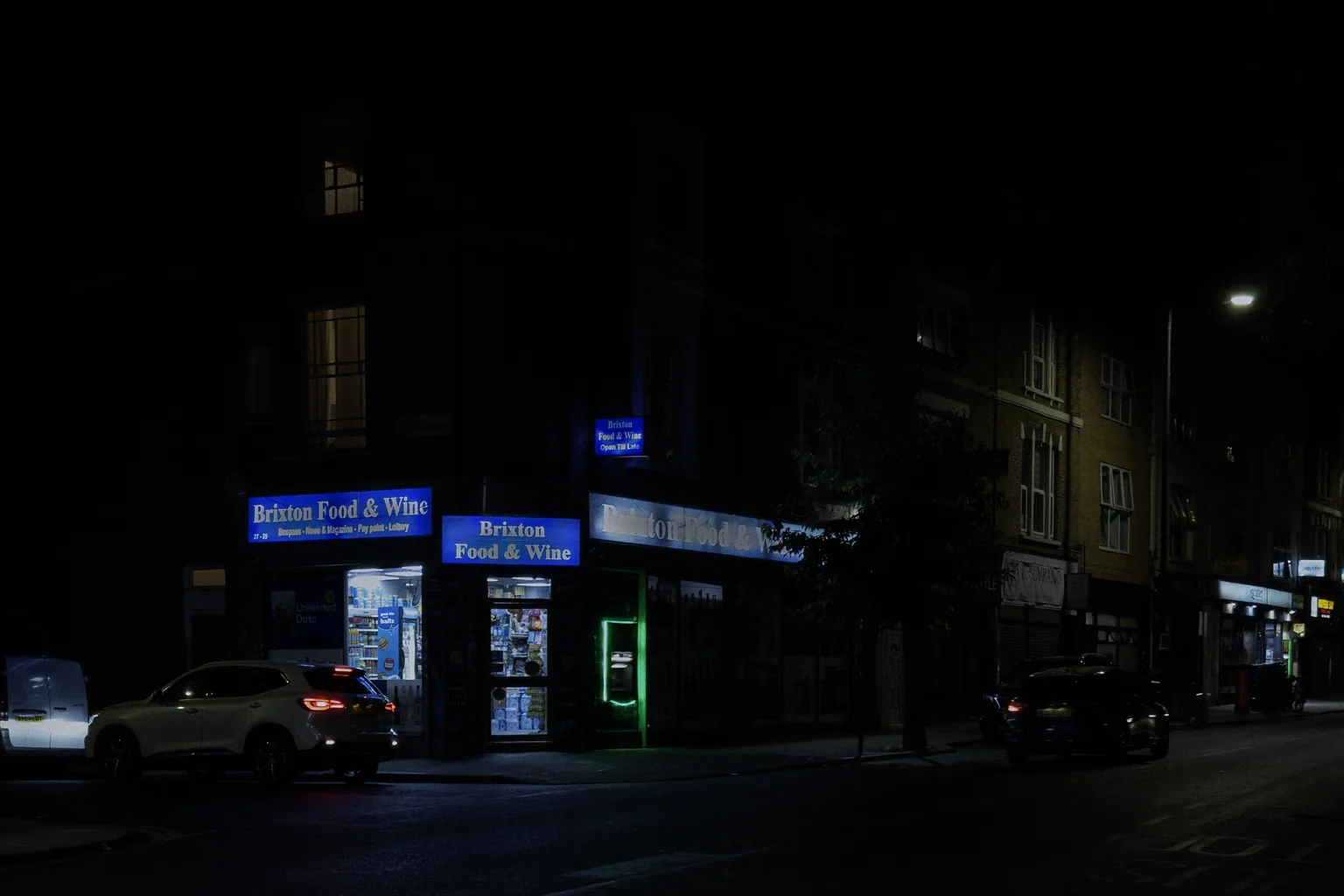 An illuminated Brixton corner shop with blue signs and a green cash machine on an otherwise dark street.