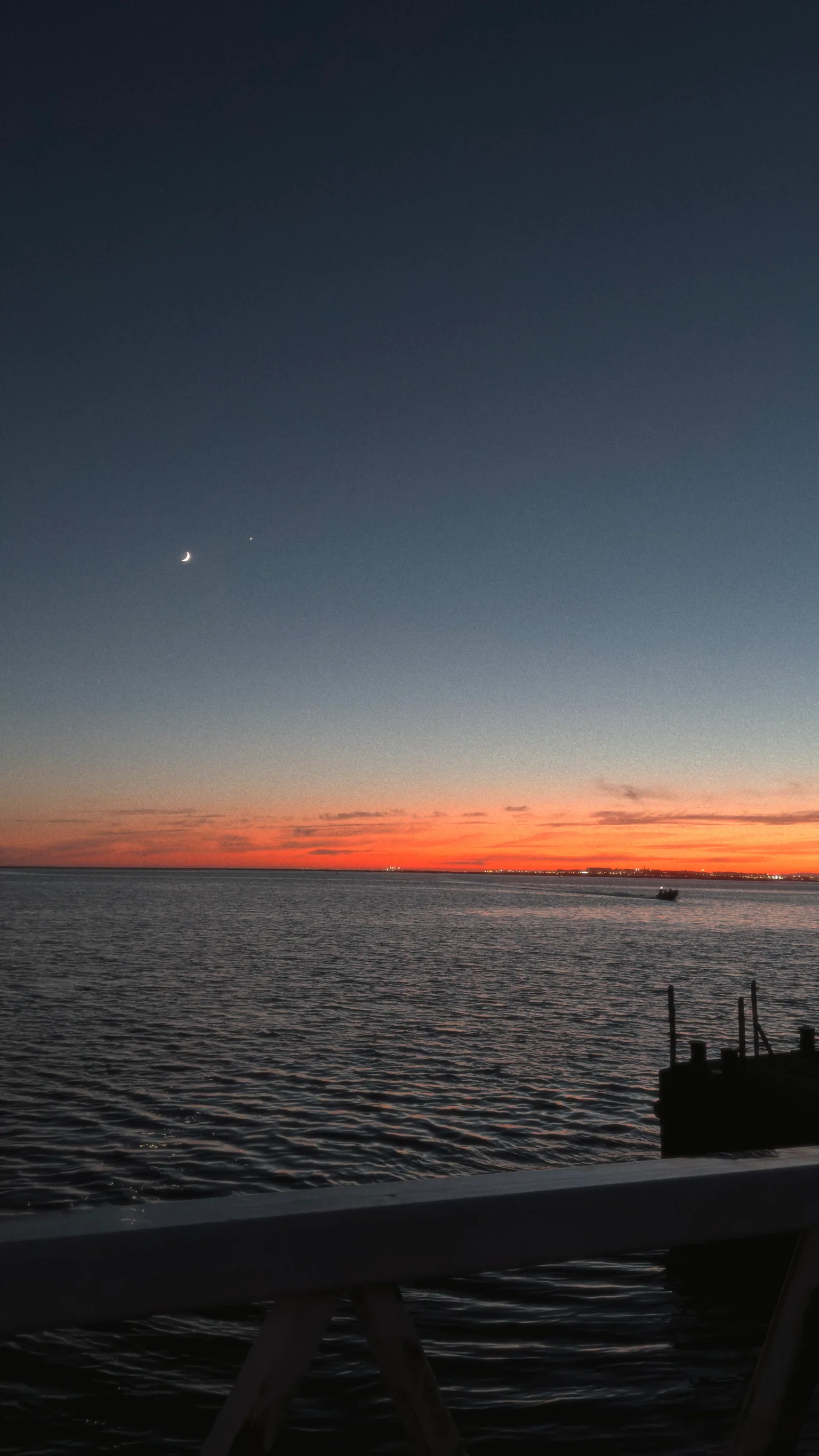 A crescent moon above darkening water and an orange horizon after sunset.