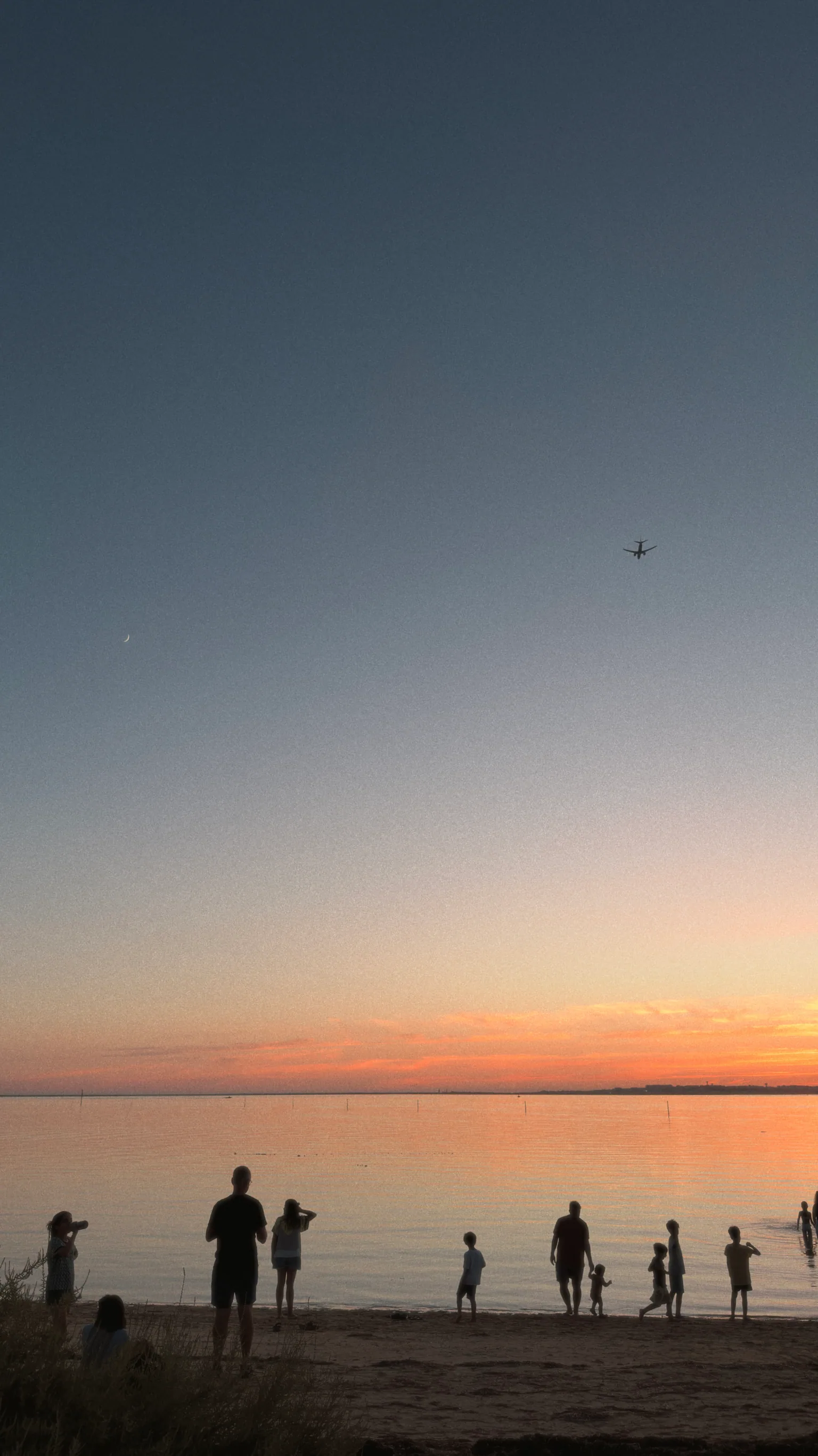 Families standing along the shoreline at sunset while a crescent moon and an aeroplane appear in the sky.
