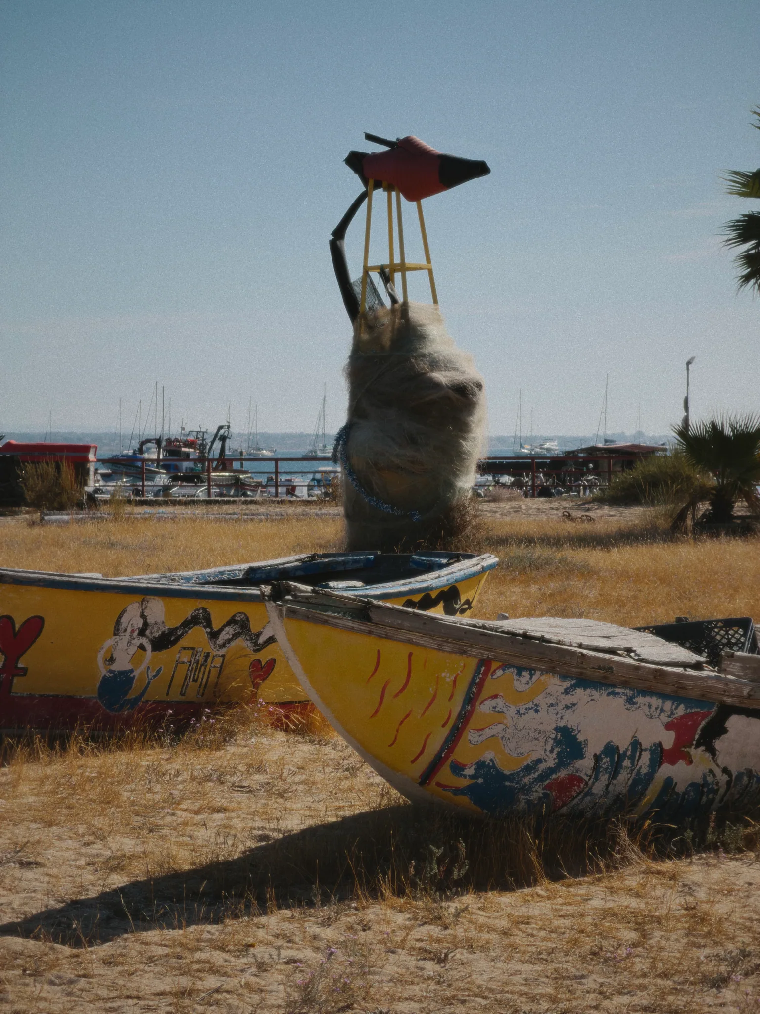 Two weathered, brightly painted fishing boats beneath a sculpture made from fishing nets near Culatra harbour.