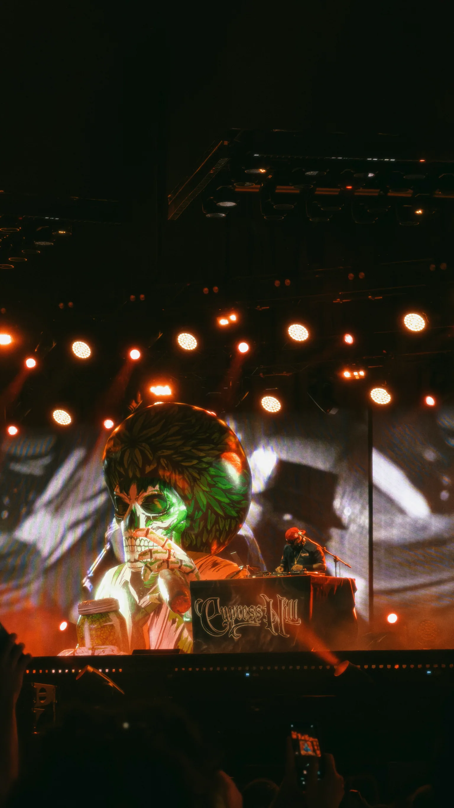 DJ Muggs performing beneath an oversized illustrated head and skull visual in orange stage light
