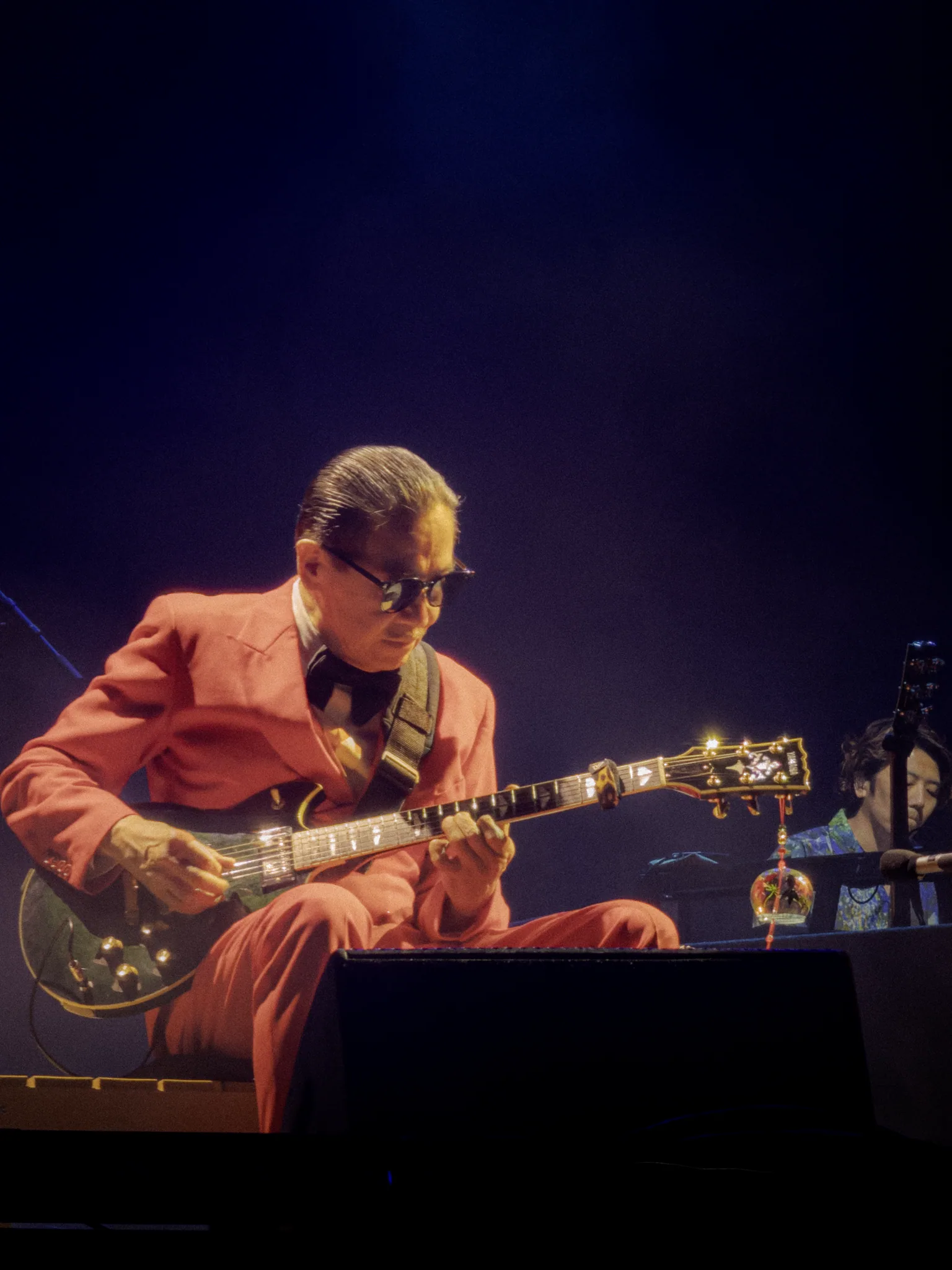 Masayoshi Takanaka seated in a red suit, playing a black guitar beneath a warm stage light.